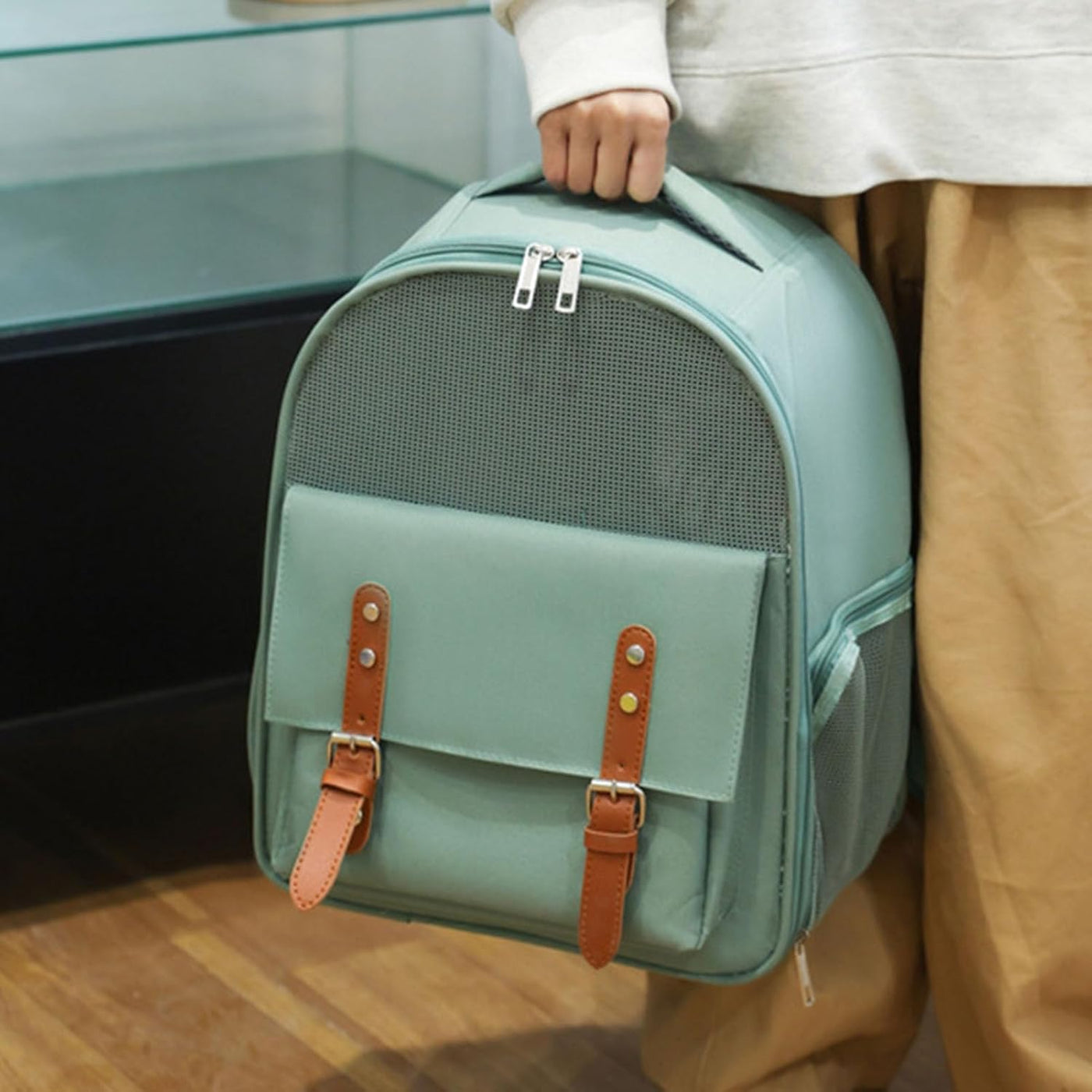 Person holding a light blue pet carrier with brown straps indoors.